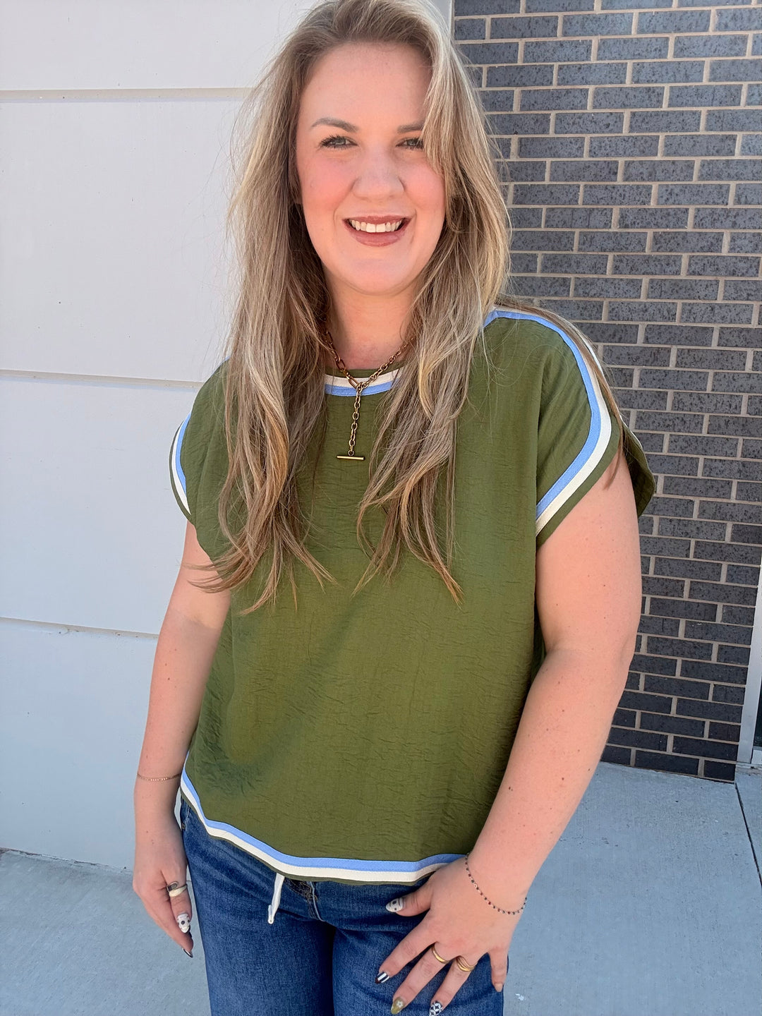 A woman wearing an olive green short-sleeve top with light blue and white striped trim on the sleeves and hem, paired with blue jeans, standing outdoors against a white wall and a dark brick wall.