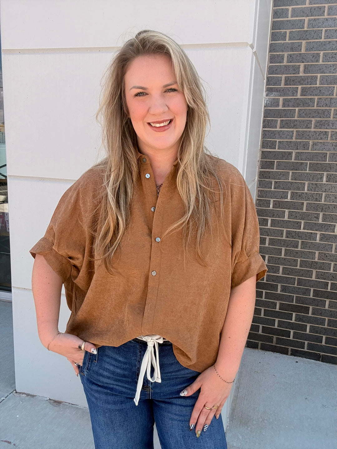 A woman with blonde hair wearing a brown short-sleeve button-up shirt and blue jeans, standing outdoors in front of a white wall and a gray brick wall.