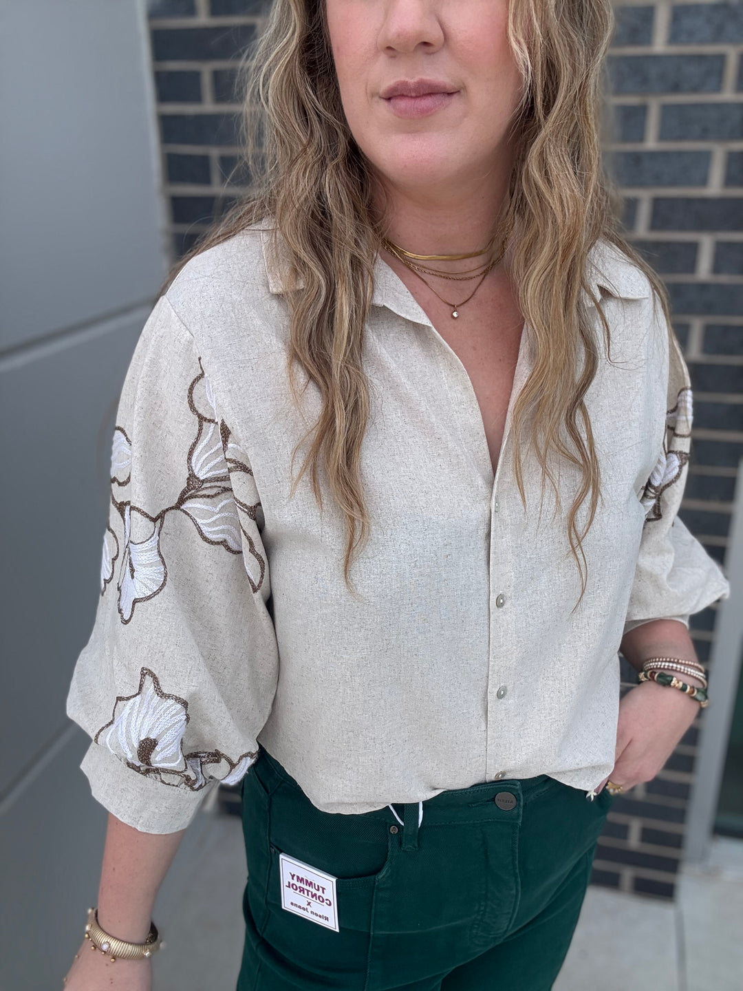 A woman wearing a light beige linen button-up shirt with brown and white floral embroidery on the sleeves, paired with dark green pants.
