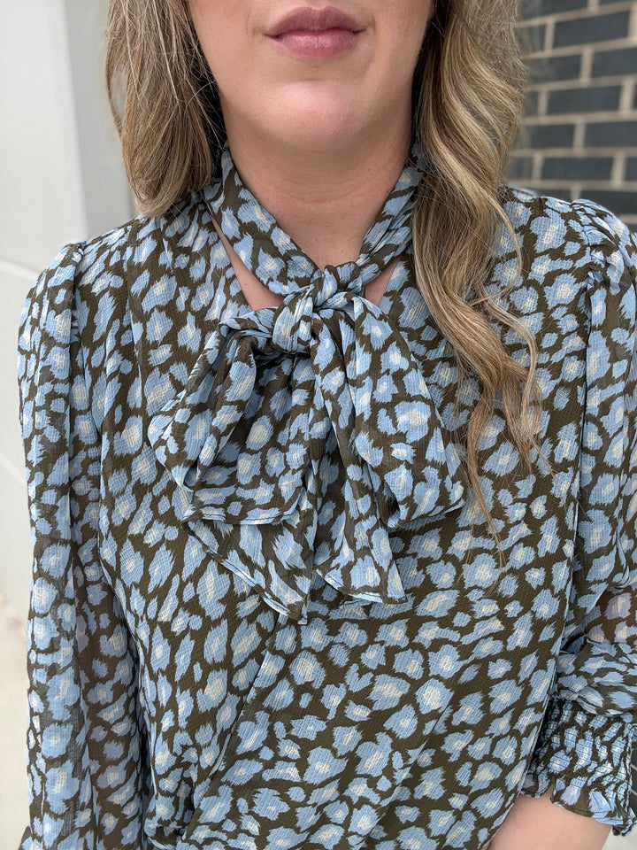 A close-up of a woman wearing a long-sleeve blouse with a blue and white abstract pattern over an olive green base, featuring a ruffled tie-neck detail.