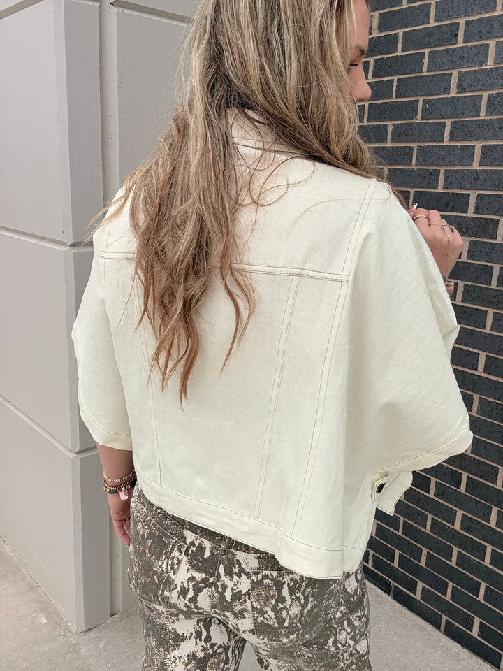 A woman viewed from behind wearing a cream-colored cropped denim jacket and camouflage pants, standing next to a gray brick wall.