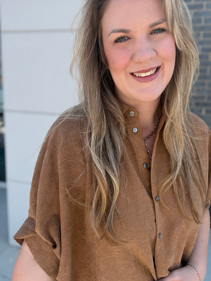 A smiling blonde woman wearing a brown short-sleeve button-up shirt, photographed in a casual outdoor setting.