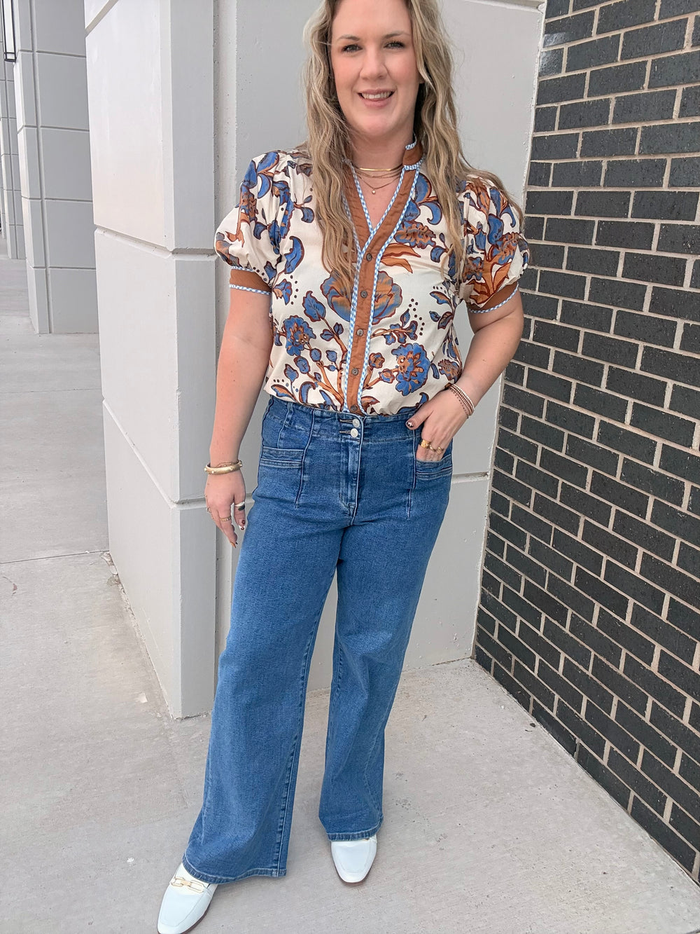 A woman modeling a short-sleeve button-front blouse with a blue, orange, and cream botanical pattern, paired with wide-leg blue jeans and white pointed shoes, standing outdoors next to a gray concrete wall and a dark brick wall.