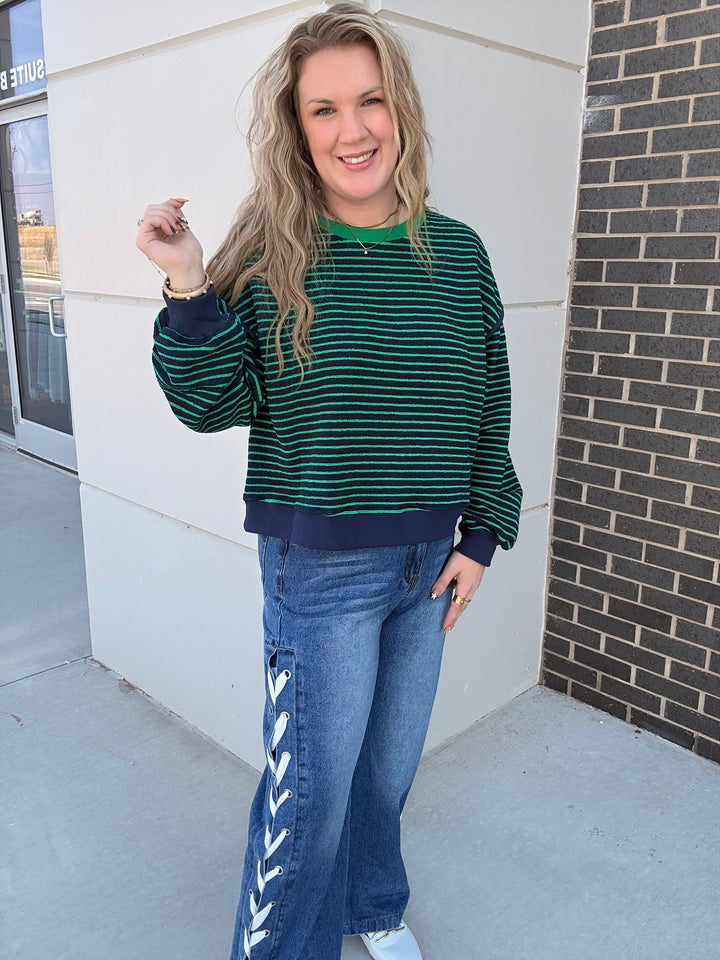 A woman wearing a green and navy striped long-sleeve crewneck sweatshirt and blue jeans with white lace-up details down the side, standing outdoors against a white wall and brick column.