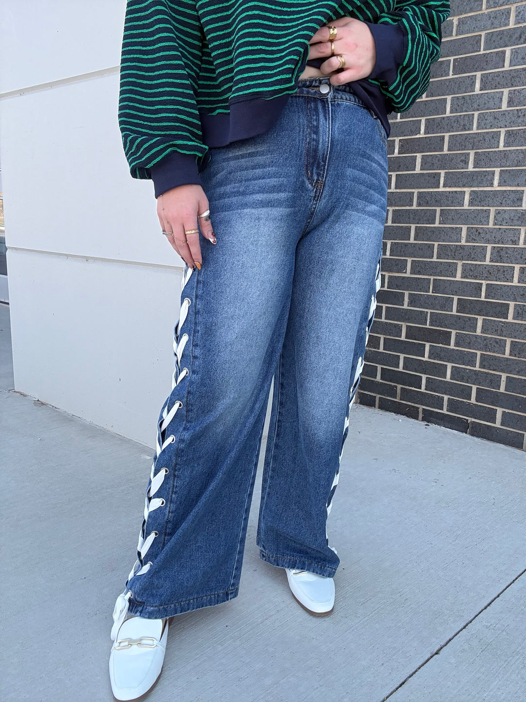 A person wearing blue wide-leg jeans with white chevron-patterned side cutouts, a green and navy striped long-sleeve top, and white loafers, standing outdoors against a white wall and a dark brick wall.