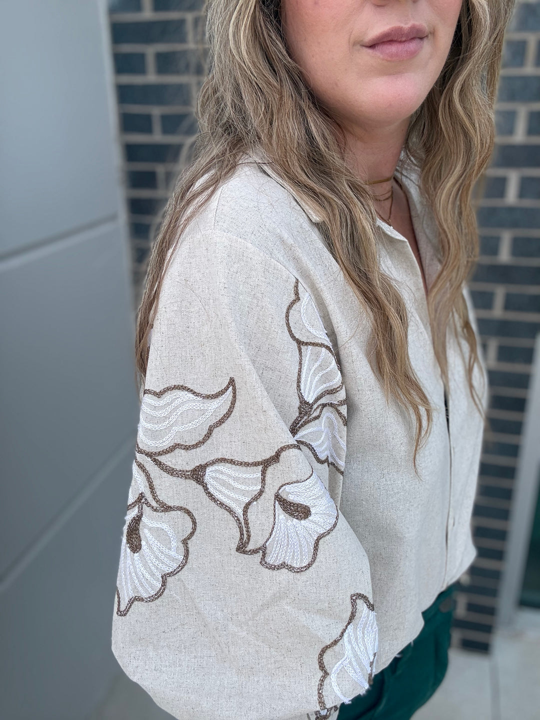 A woman wearing a beige linen button-up shirt with white and brown floral embroidery on the sleeves, paired with a dark green top underneath.