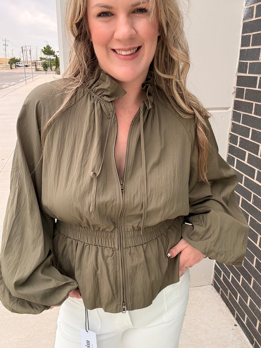 A woman wearing an olive green lightweight zip-up jacket with a high drawstring collar and elasticized waist, paired with white pants, standing outdoors next to a brick wall.