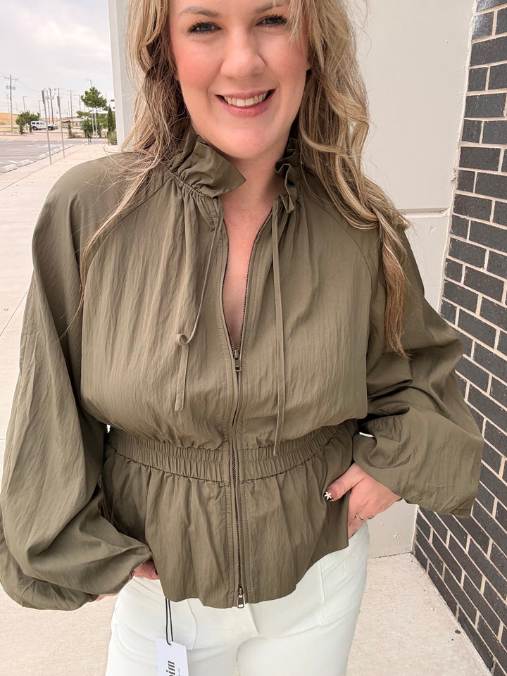 A woman wearing an olive green lightweight zip-up jacket with a high drawstring collar and elasticized waist, paired with white pants, standing outdoors next to a brick wall.