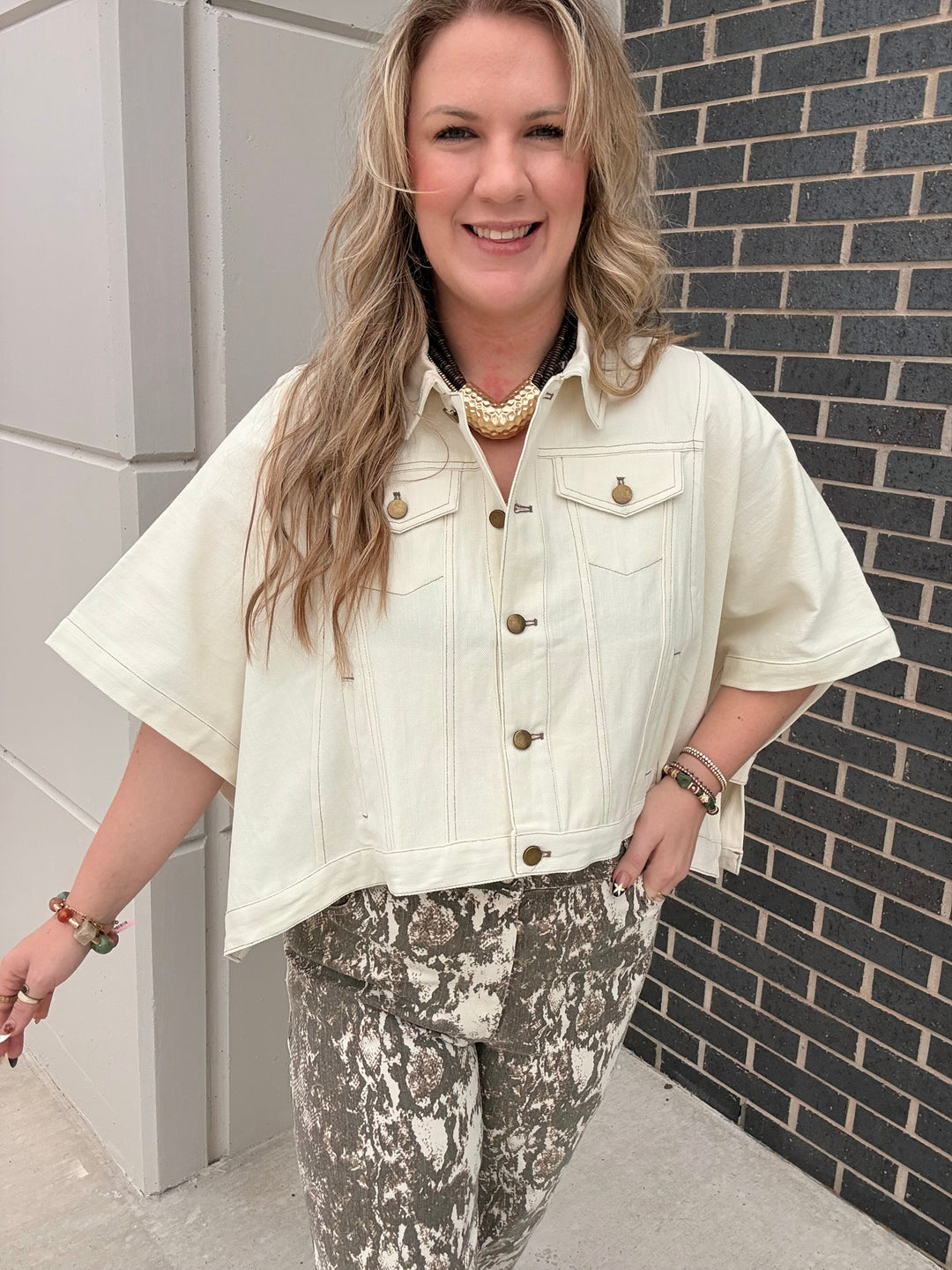 A woman wearing a cream-colored short-sleeve denim jacket with gold-tone buttons and a matching patterned pair of pants, standing outdoors against a gray brick wall.