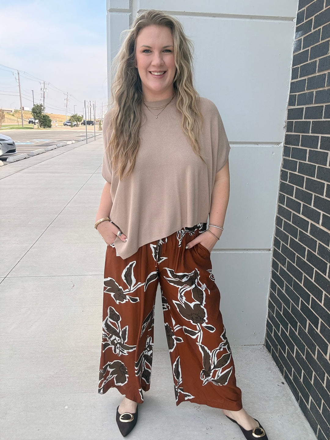 A woman stands outdoors on a sidewalk wearing a beige short-sleeve top and brown wide-leg pants with a white floral pattern, paired with black pointed flats.