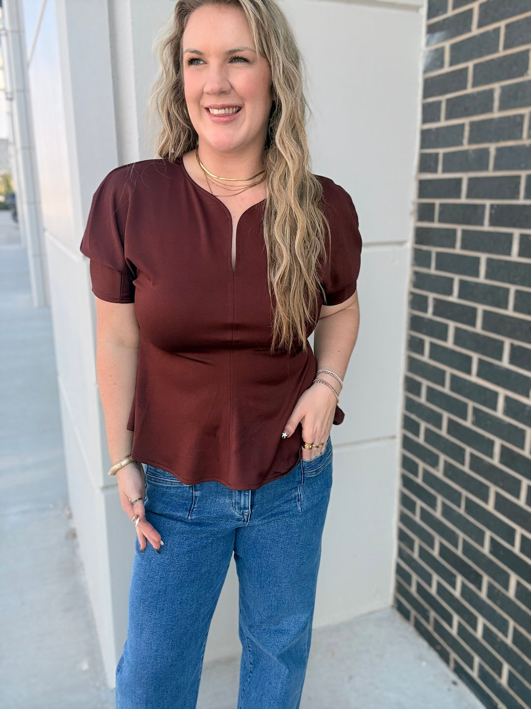A woman wearing a short-sleeve maroon top with a split neckline and blue jeans, standing outdoors next to a white wall and a dark brick column.
