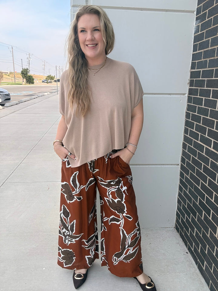A woman stands outdoors wearing a beige short-sleeve top and rust-brown wide-leg pants with a white floral pattern, paired with black pointed flats.