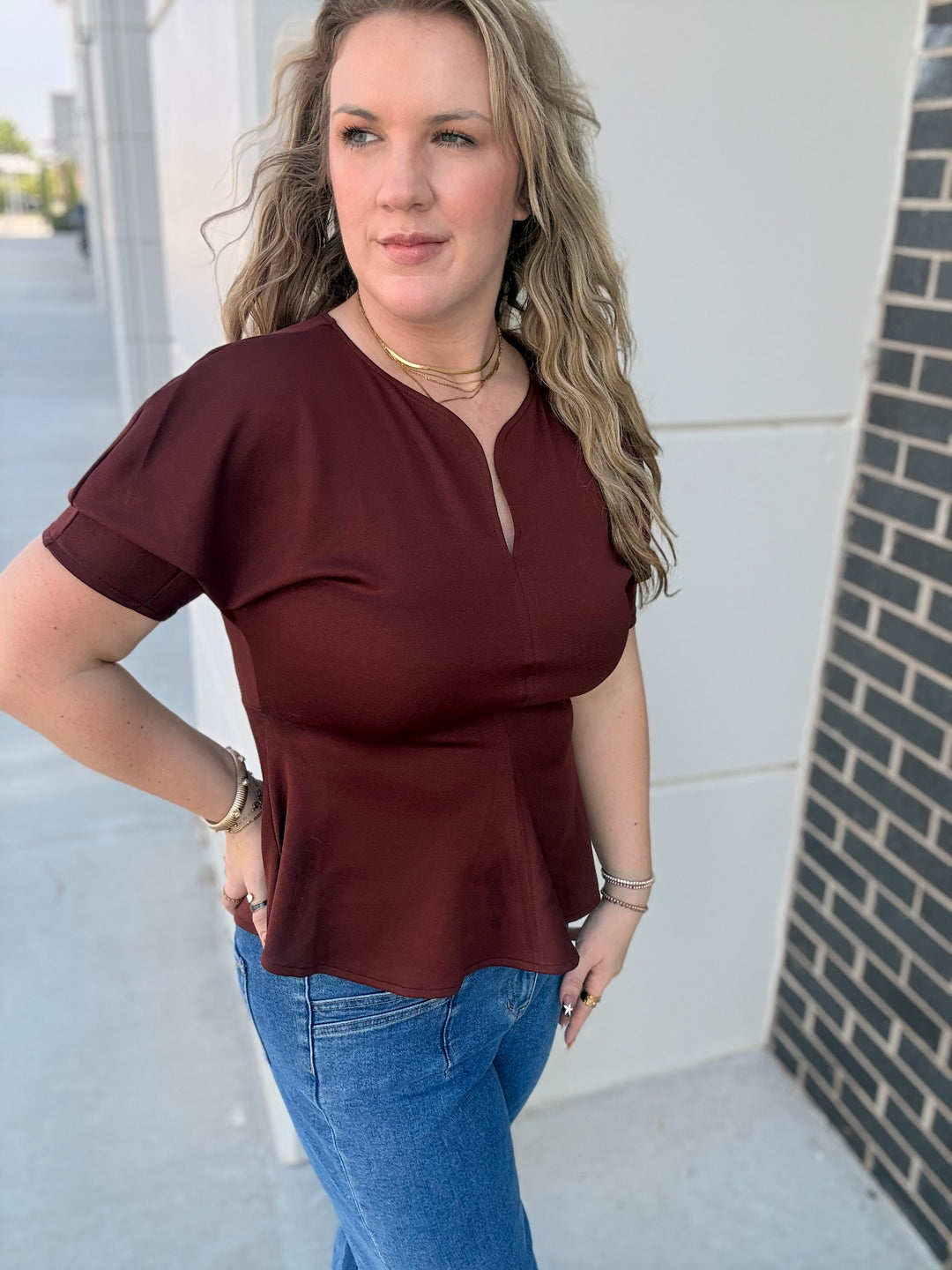 A woman wearing a short-sleeve maroon top with a split neckline and blue jeans, standing outdoors against a light-colored building wall.