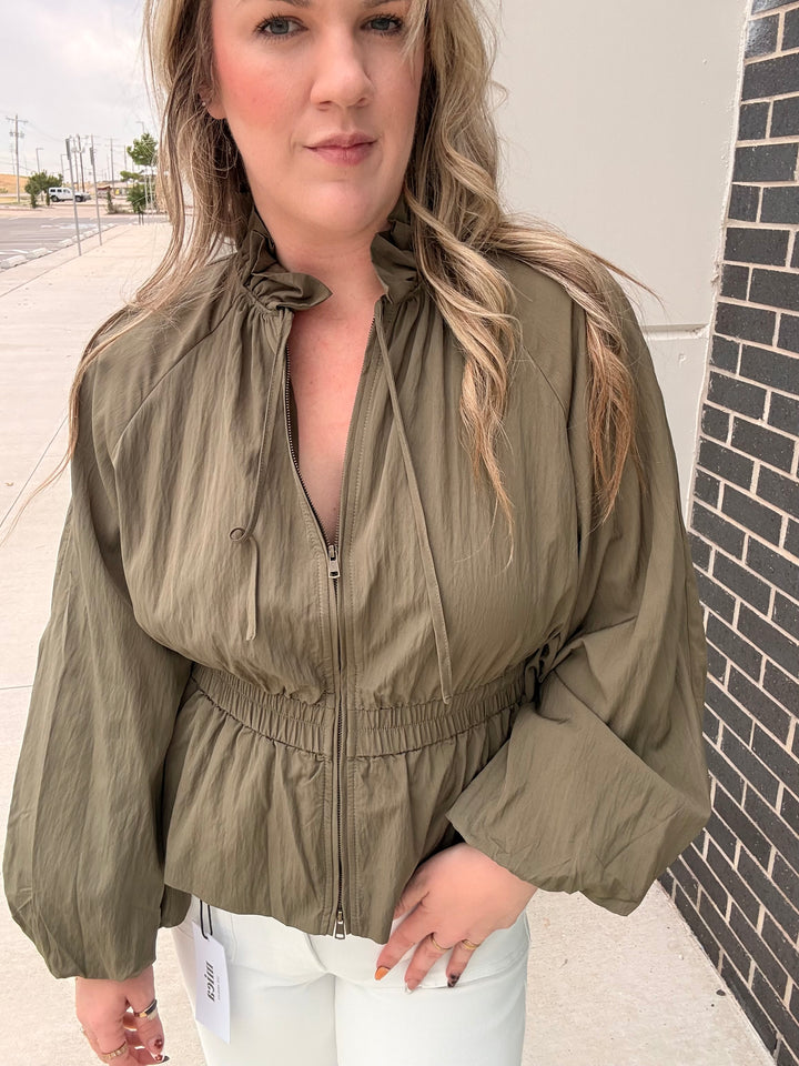 A woman wearing an olive green zip-up jacket with a gathered waist and white pants, standing outdoors next to a brick wall.