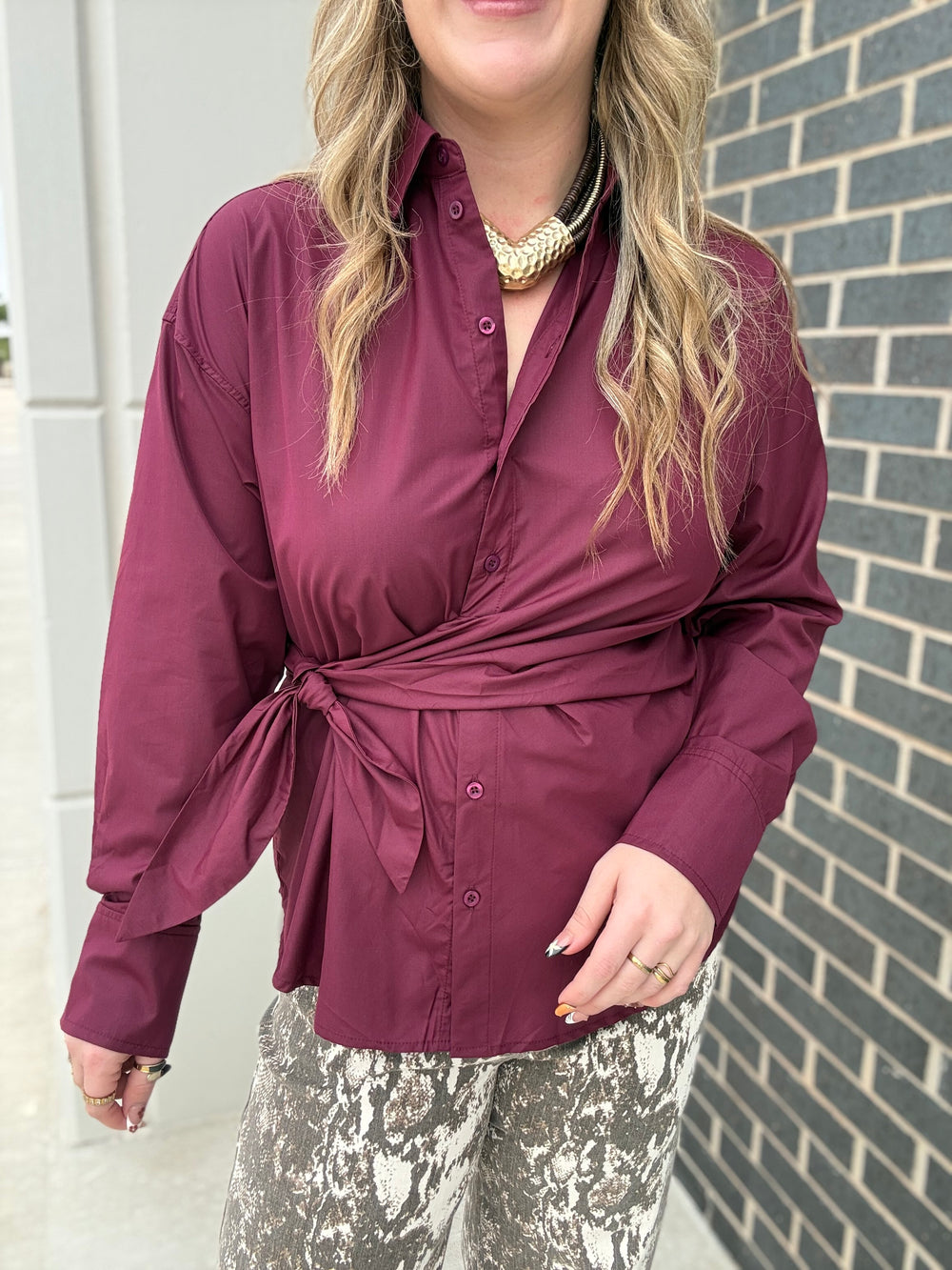 A woman wearing a long-sleeve wrap-style button-up blouse in deep burgundy, paired with gray and white patterned pants, standing outdoors next to a gray brick wall.