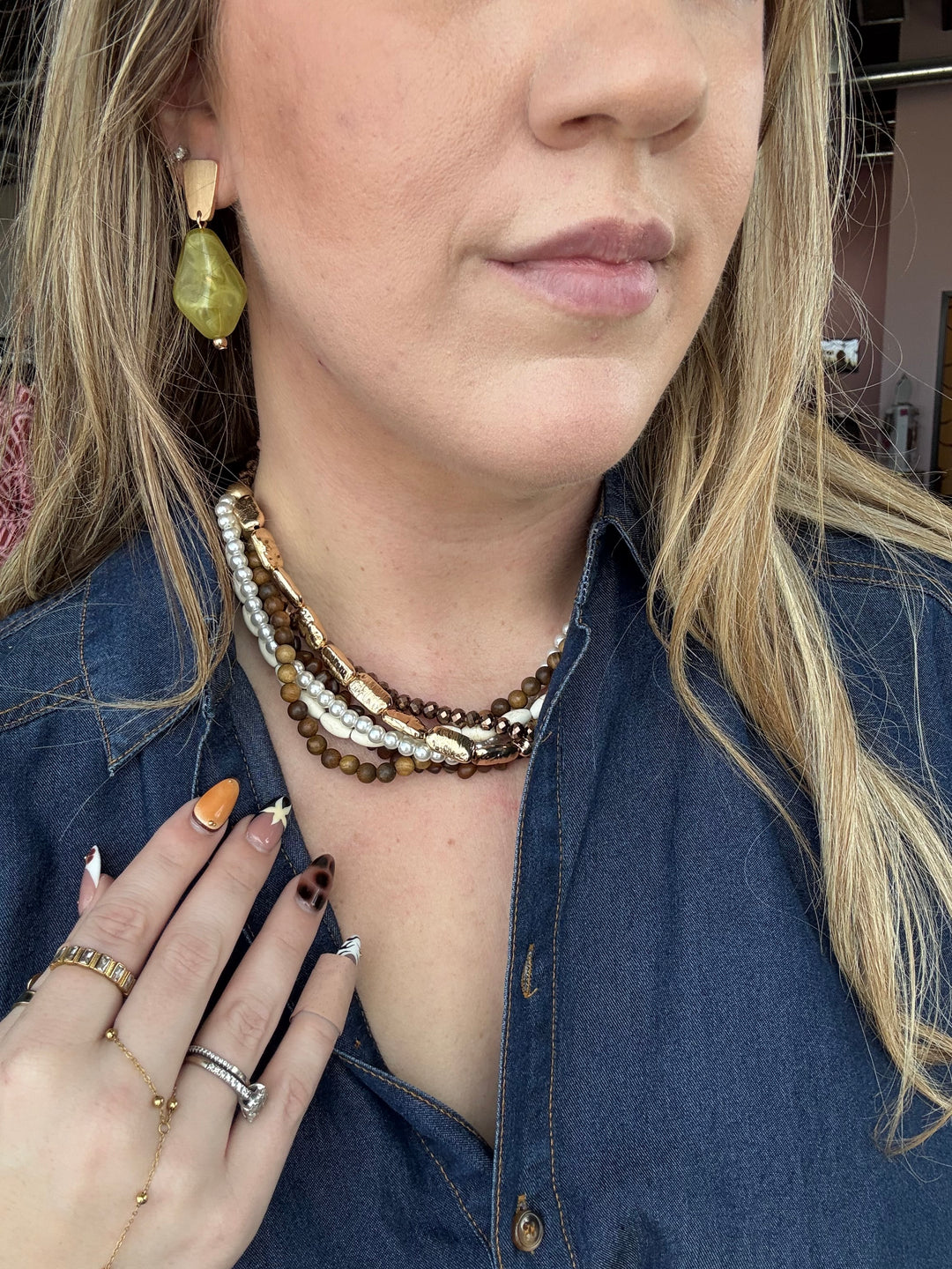 A close-up lifestyle shot of a woman wearing a multi-strand beaded necklace with brown, cream, and gold beads, along with matching rings and a green teardrop earring.