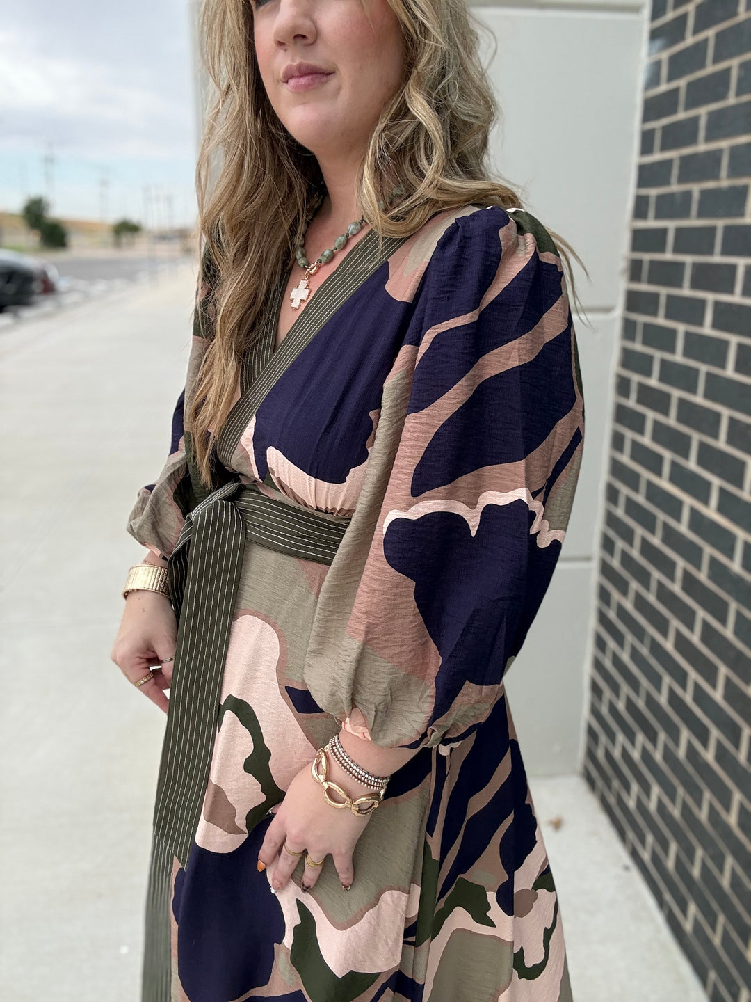 A woman wearing a long-sleeve wrap dress with a navy, beige, and olive green abstract pattern, standing outdoors next to a brick wall.