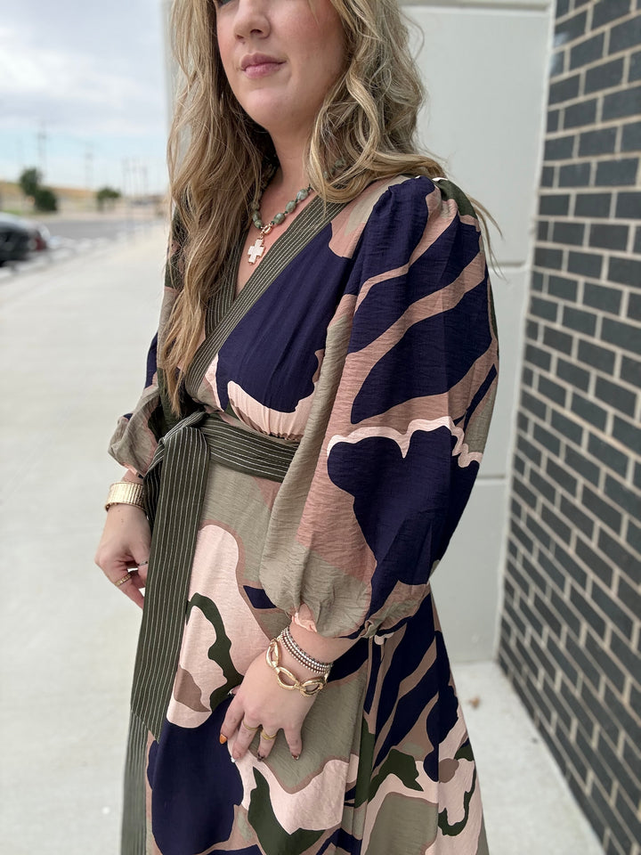 A woman wearing a long-sleeve wrap dress with a navy, beige, and olive green abstract pattern, standing outdoors next to a brick wall.
