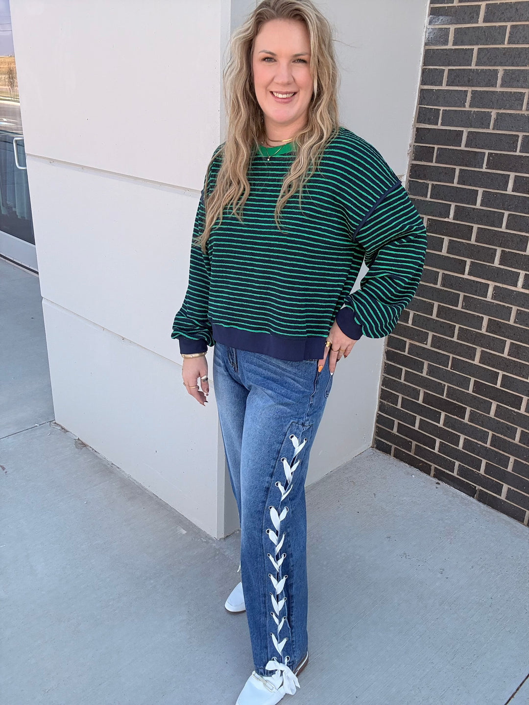 A woman modeling a dark green and navy striped crewneck sweatshirt with blue jeans featuring white lace-up detailing down the side, paired with white sneakers, standing outdoors against a modern building wall.