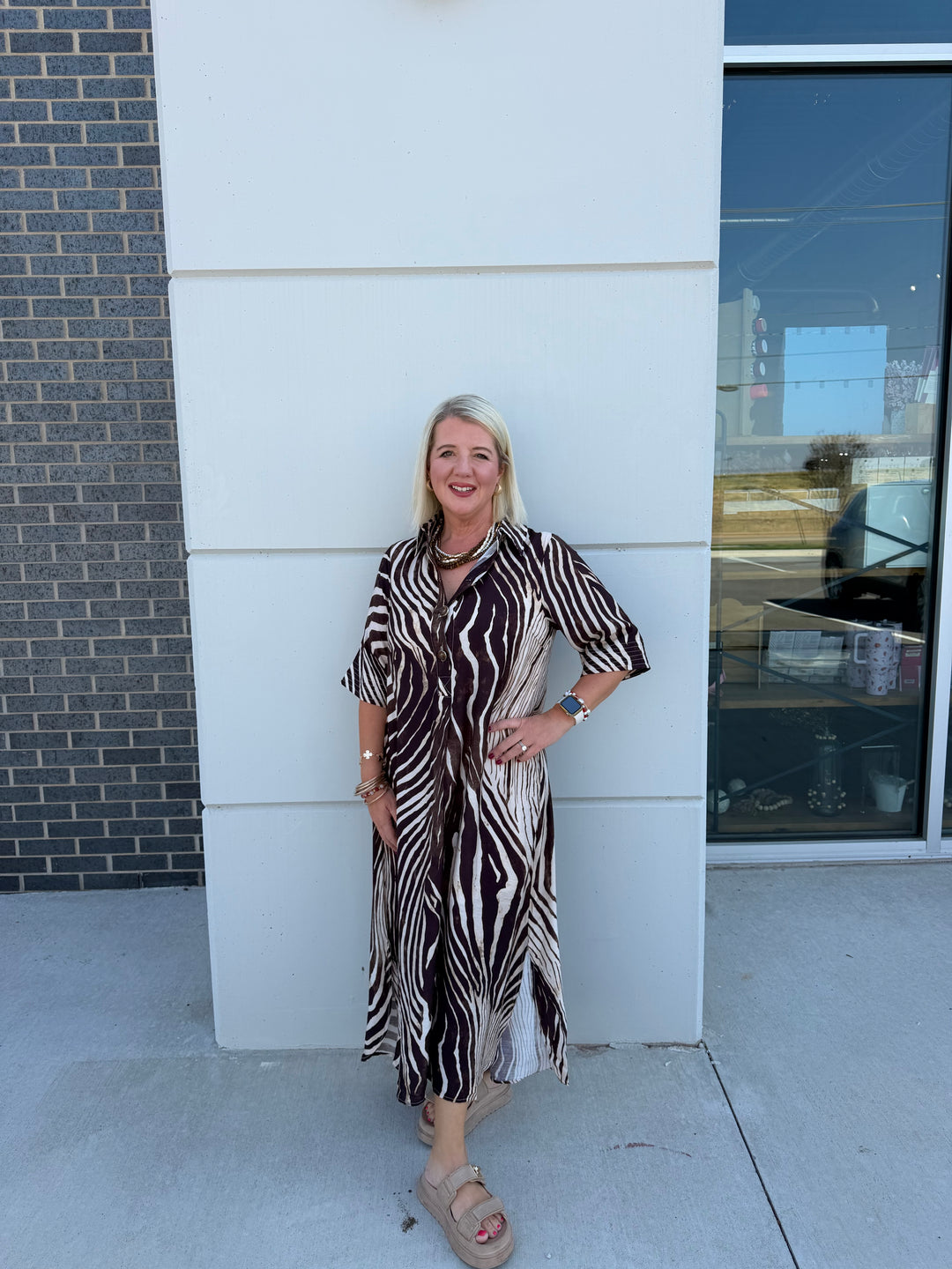 A woman with blonde hair stands outdoors against a white building wall, wearing a brown and beige zebra-print maxi dress with short wide sleeves and a tie waist, paired with beige platform sandals.