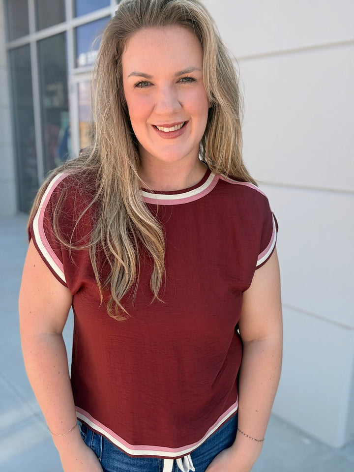 A smiling blonde woman wearing a short-sleeve crewneck top in burgundy with cream and white striped trim, paired with blue jeans, standing outdoors against a light-colored building wall.