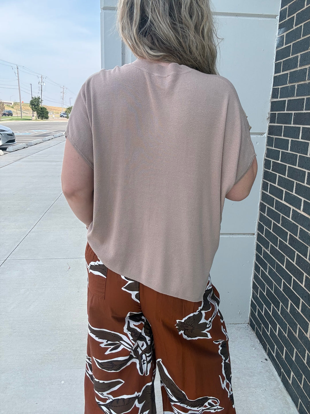 A person shown from behind wearing a loose, short-sleeve dusty pink top and brown wide-leg pants with a white floral pattern, standing outdoors next to a white wall and a dark brick column.