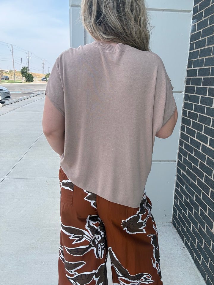 A person shown from behind wearing a loose, short-sleeve dusty pink top and brown wide-leg pants with a white floral pattern, standing outdoors next to a white wall and a dark brick column.