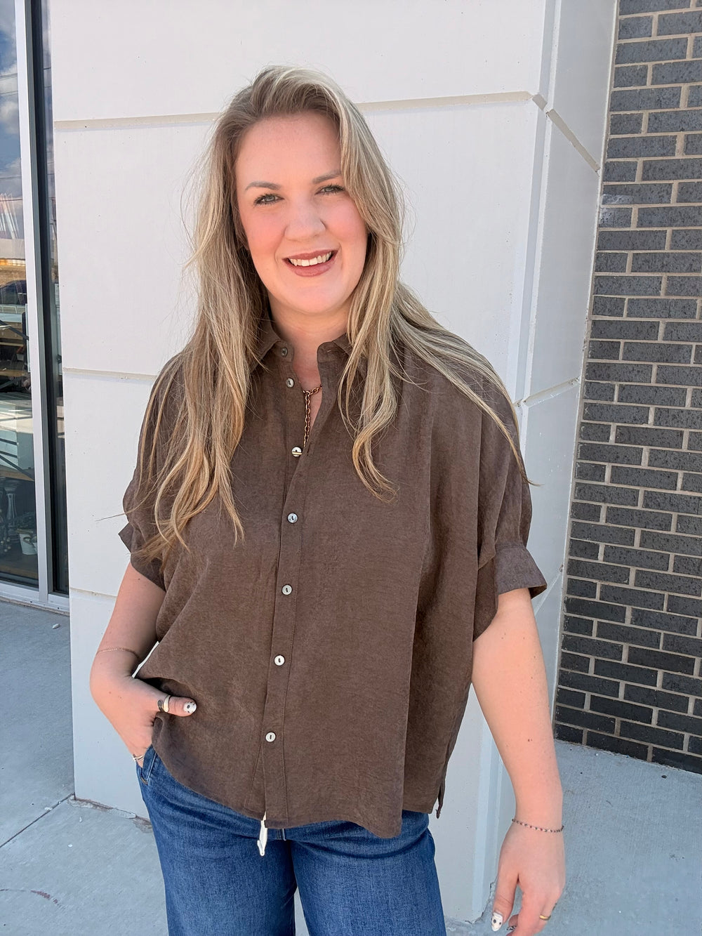 A woman with blonde hair wearing a brown short-sleeve button-up shirt and blue jeans, standing outdoors in front of a modern building wall.