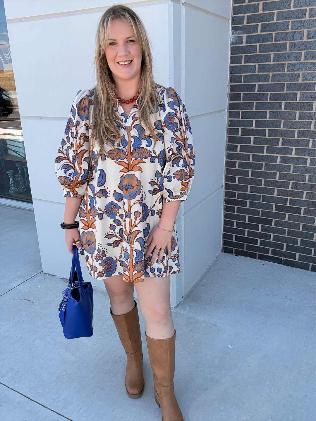 A woman stands outdoors wearing a short-sleeve cream dress with a blue and brown floral pattern, paired with tan knee-high boots and holding a bright blue handbag.
