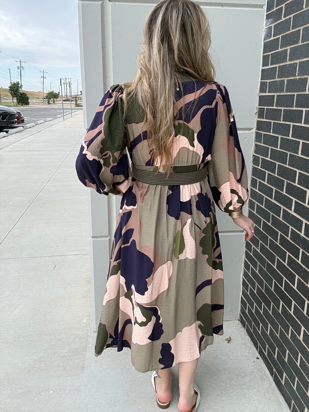 A woman viewed from behind wearing a long-sleeve midi dress with a camouflage pattern in olive green, navy blue, and dusty pink, featuring a matching fabric belt at the waist. She is standing outdoors next to a gray utility box and a dark brick wall.