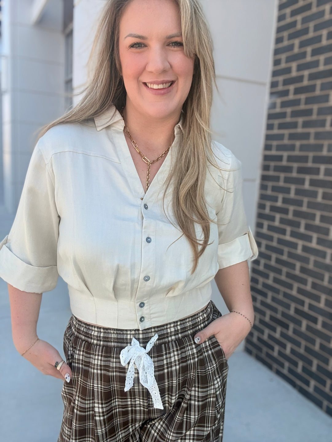 A woman wearing a cream-colored short-sleeve button-up shirt and brown plaid pants, standing outdoors against a brick wall background.