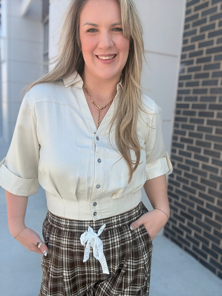 A woman wearing a cream-colored short-sleeve button-up shirt and brown plaid pants, standing outdoors against a brick wall background.