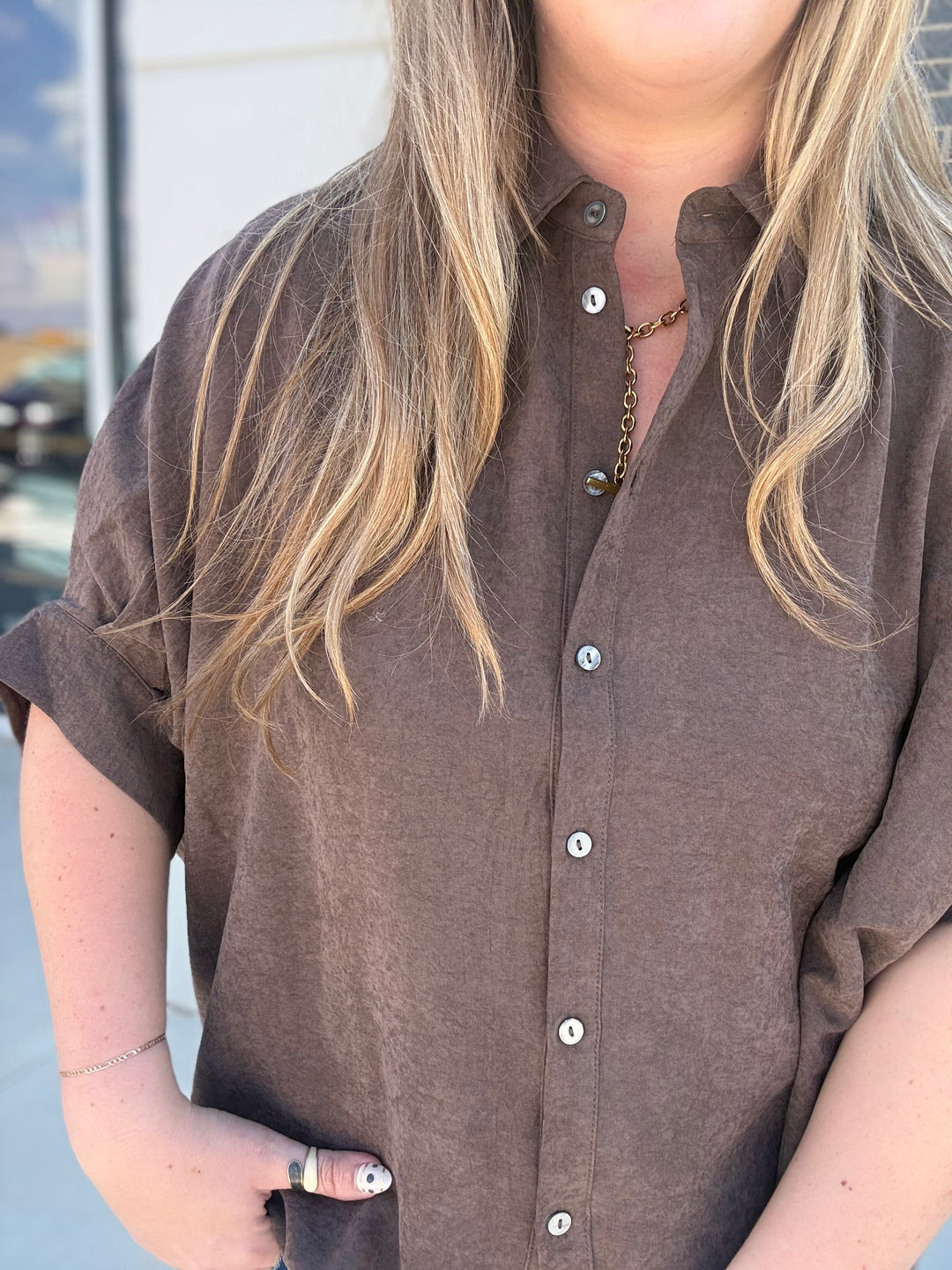 A close-up of a person wearing a brown short-sleeve button-up shirt with rolled cuffs and silver buttons, standing outdoors.