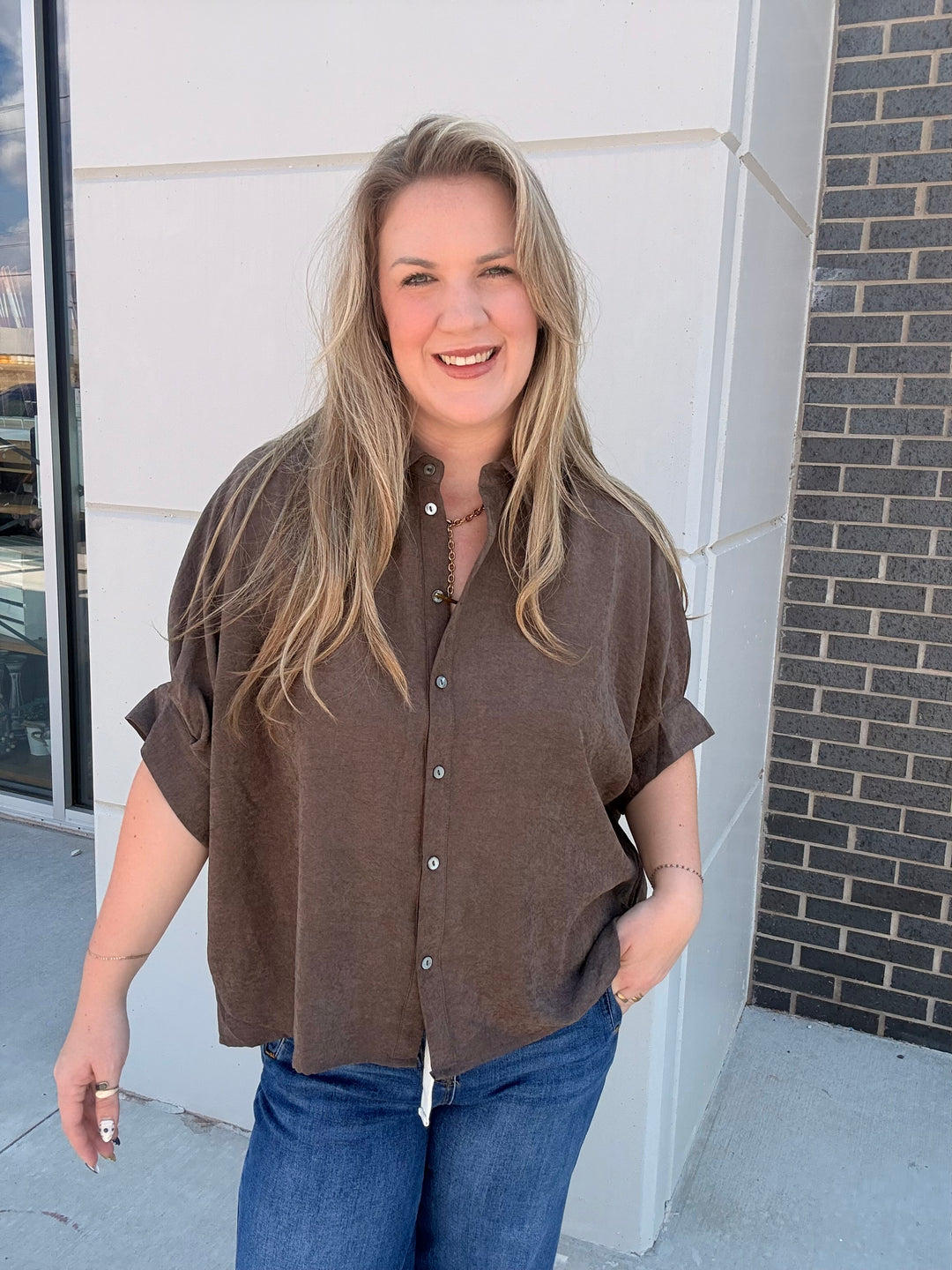A woman with blonde hair wearing a brown short-sleeve button-up shirt and blue jeans, standing outdoors in front of a modern building with a white wall and dark brick column.