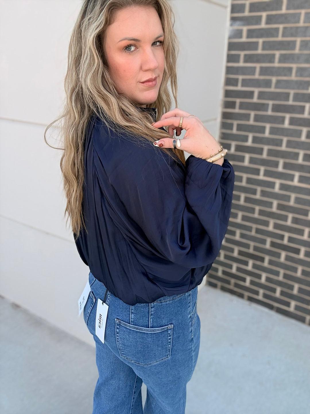 A woman wearing a navy blue long-sleeve blouse and blue jeans, photographed outdoors against a white wall and brick wall background.