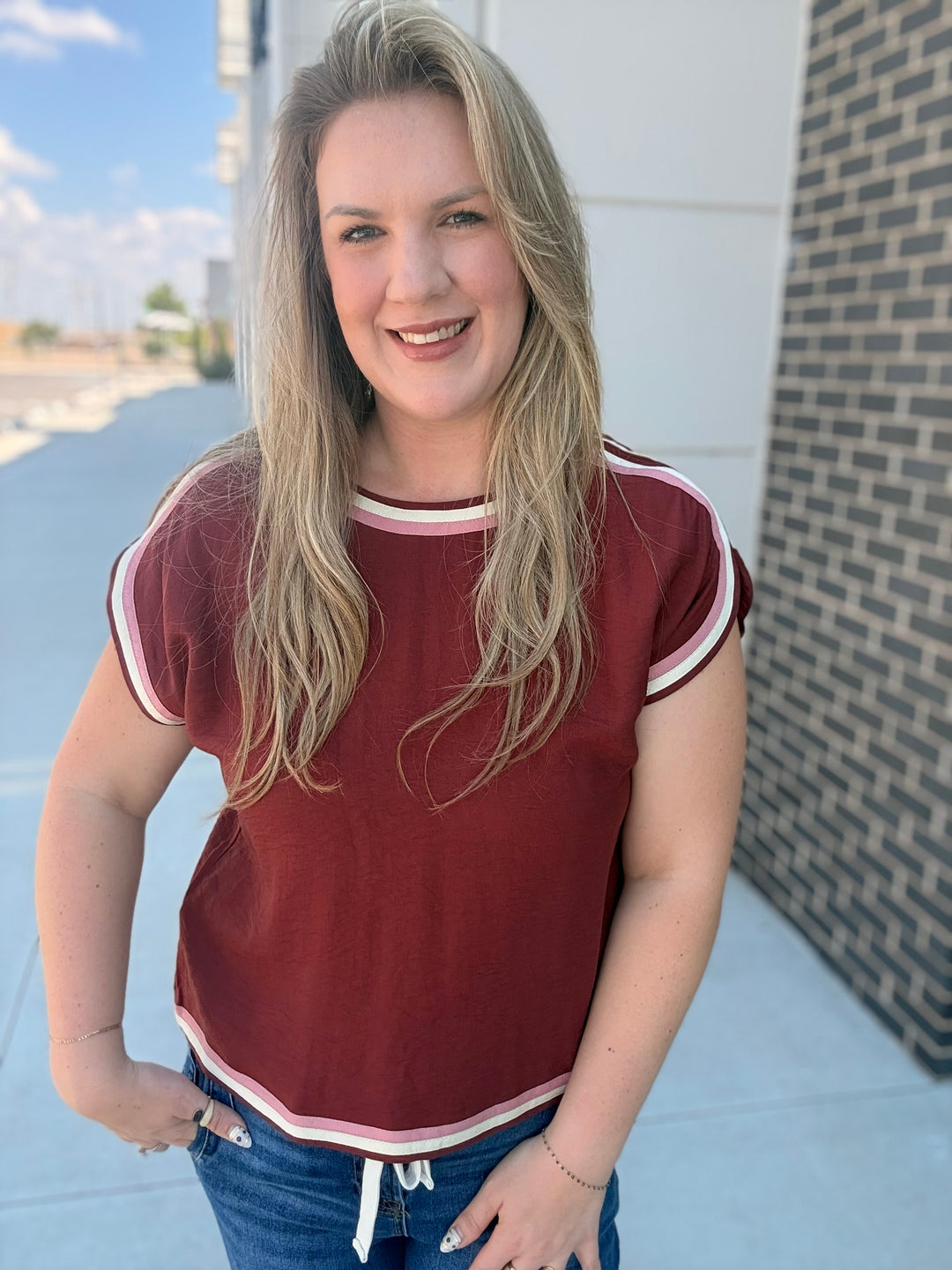 A smiling blonde woman wearing a short-sleeve crewneck t-shirt in maroon with white and light pink striped trim, paired with blue jeans, standing outdoors next to a brick wall.
