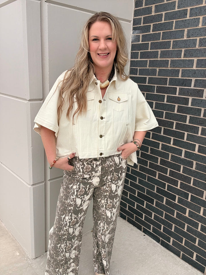 A woman stands outdoors wearing a cream short-sleeve button-up shirt and gray-and-white abstract print pants, posing in front of a gray wall and a black brick wall.