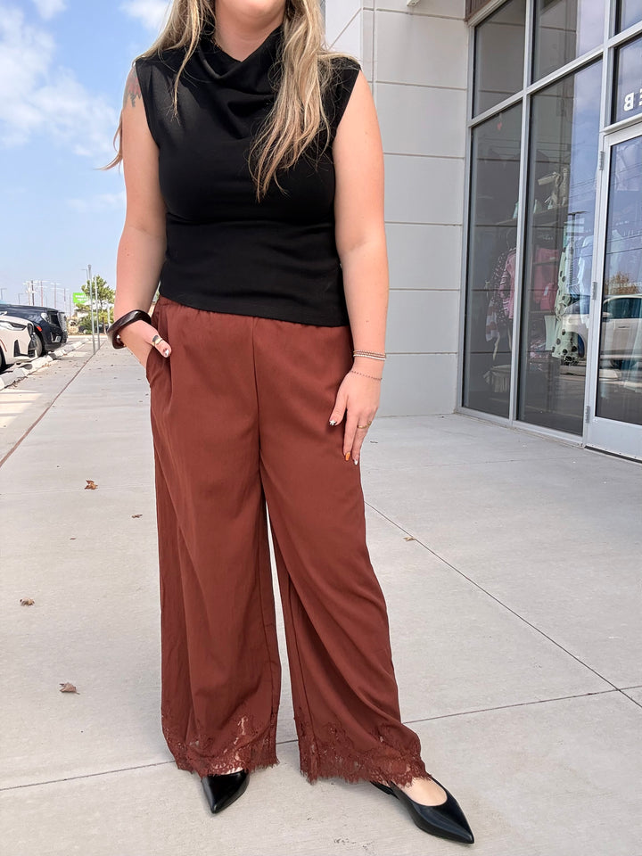 A woman wearing a black sleeveless cowl-neck top and rust-brown wide-leg pants with lace trim at the hems, standing on a sidewalk in front of a modern building.