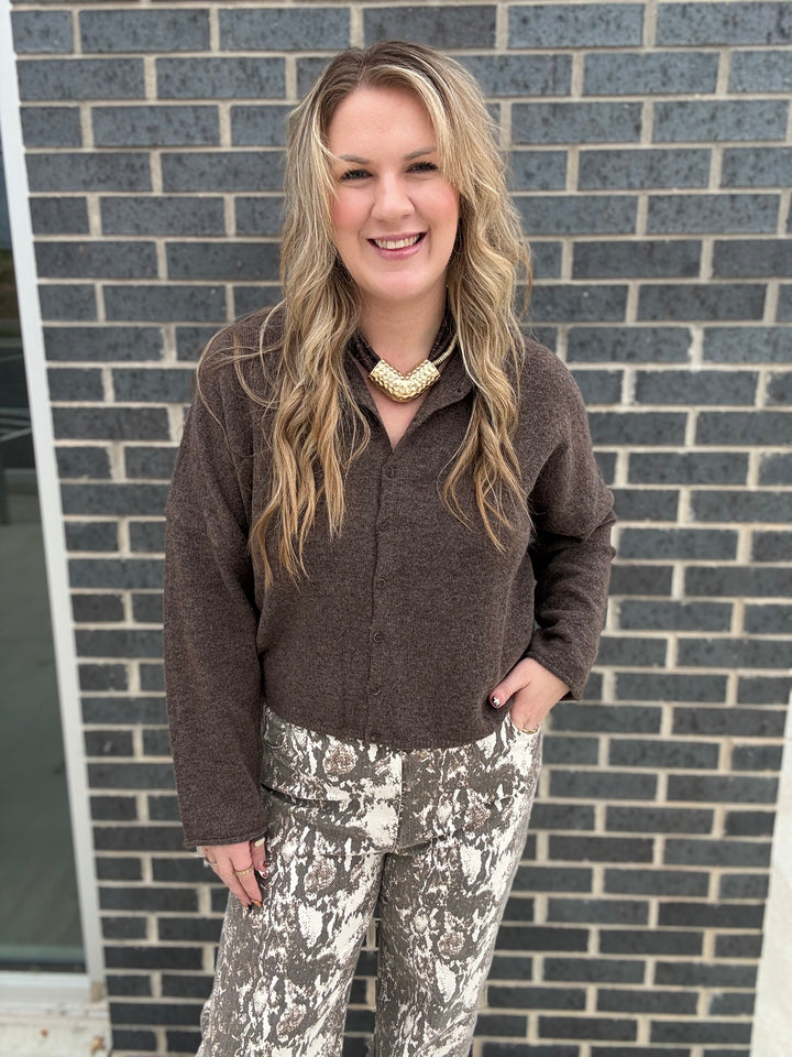A woman with blonde hair stands in front of a gray brick wall, wearing a brown fuzzy long-sleeve button-up top and brown-and-white patterned pants.