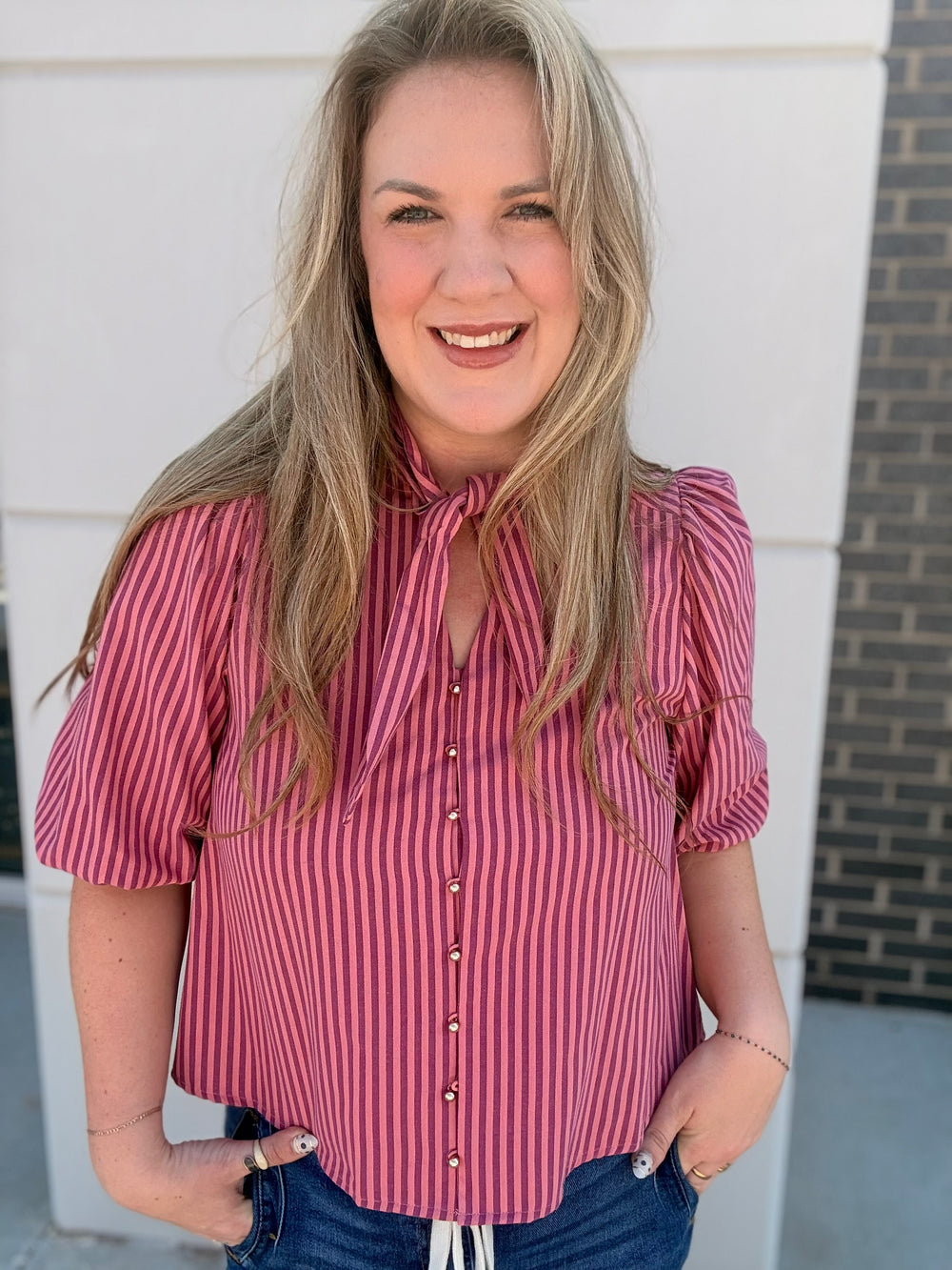 A smiling blonde woman wearing a pink and red vertically striped short-sleeve blouse with a tie-neck detail and puff sleeves, paired with blue jeans, standing outdoors against a white wall.