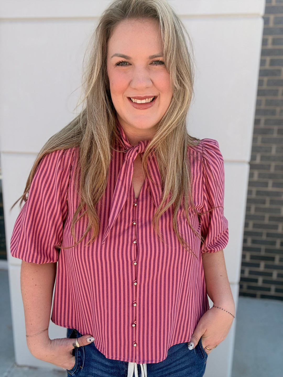 A smiling blonde woman wearing a pink and red vertically striped short-sleeve blouse with a tie-neck detail and puff sleeves, paired with blue jeans, standing outdoors against a white wall.