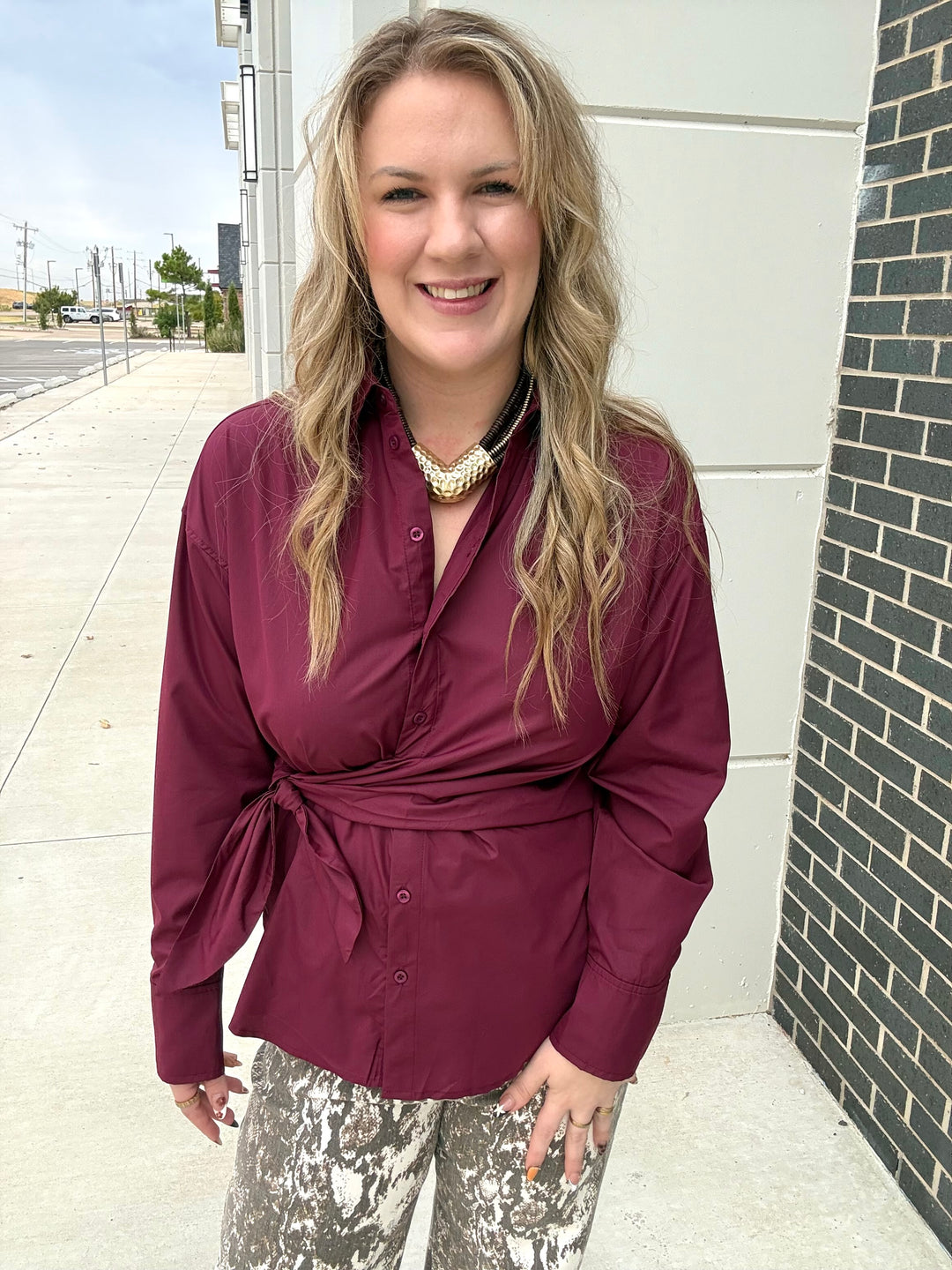 A woman with blonde hair stands outdoors wearing a long-sleeve wrap-style blouse in deep burgundy with a tie waist, paired with patterned pants and a chunky gold necklace.