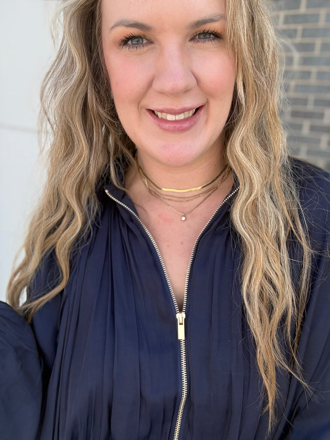 A smiling woman with long wavy blonde hair wearing a navy blue zip-up jacket and layered gold necklaces, photographed in a close-up portrait.